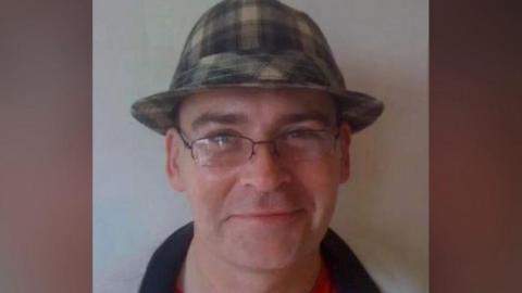 A closeup of a middle-aged man smiling, wearing glasses and green checkered bucket hat and red t-shirt and grey tracksuit jacket