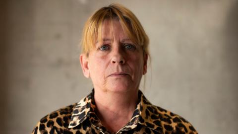 Martina Dillon - a woman with blonde hair wearing a leopard print button-up shirt stares at the camera. She is standing in front of a grey wall which has been blurred in the background.