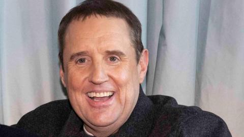 Peter Kay smiles at the camera with short greying hair and wearing a grey jacket