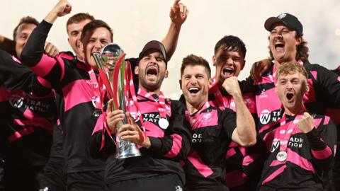 Somerset players wearing pink and black shirts lift the T20 Blast trophy and celebrate