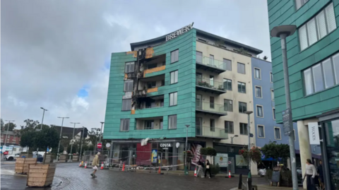 A photo of the building in the aftermath of the fire in the town square. The block of flats is partially blackened by the smoke. The building has been cordoned off and there are orange and white cones in front of it. Some people are walking nearby. It is cloudy.