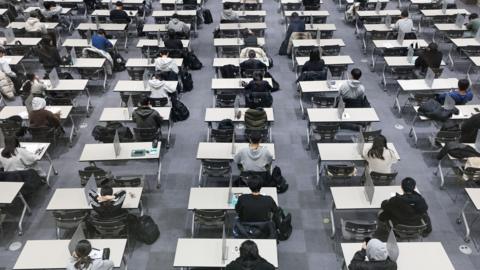 Students sat in rows on desks about to take an exam. Grey flooring.