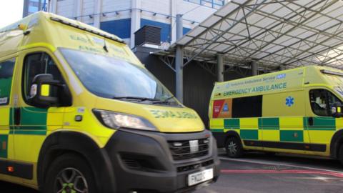 Two yellow emergency ambulance vans are parked outside a beige building with metal bars and a white cover going across it.
