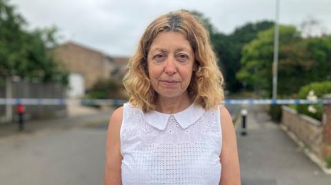 Frances Pulley a brown haired woman wearing a white top and stood in a street with a police cordon behind her.