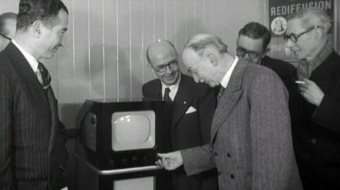 Men standing around a television set.