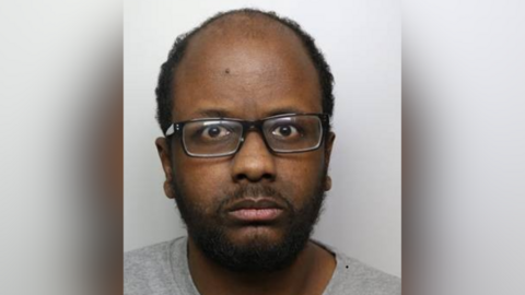 A mugshot of Zakaria Mohammed, who is wearing a grey T-shirt and black-rimmed glasses.