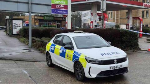 An Essex Police car. It is white with police written in blue letters on its bonnet and a blue and yellow stripe on its side with Essex Police on its door. It is parked at an entrance/exit to an Esso  petrol station in Harlow.