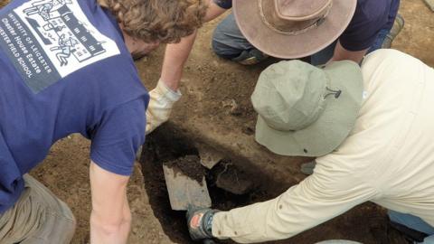 A figure in a beige shirt and olive hat shovels earth out of a hole while two men in navy T-shirts crouch close by looking on.