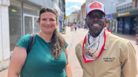 Sarah Tieck and Raph Adom standing in a street. Sarah is wearing a green top, Raph is wearing a cream coat, a neck scarf and a trucker's cap