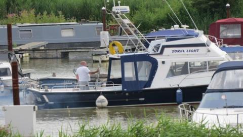 A white and blue small boat, called Sorel Light, is on a river, with greenery in the background. A man is standing on the boat with his back to the camera.