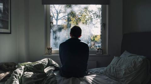 A man looks out of a window. He is in a dark room on an unmade bed. The atmosphere seems negative. 