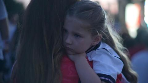 A young blonde girl wearing a white England top clings on to a woman who is wearing a red England top.