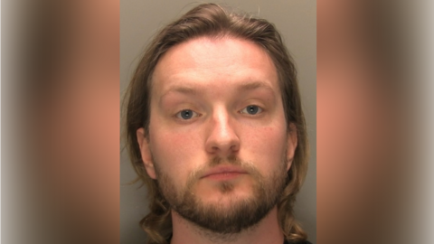 A police custody shot of Rhys Lynn. He has long brown hair and brown facial hair. The wall behind him is grey.