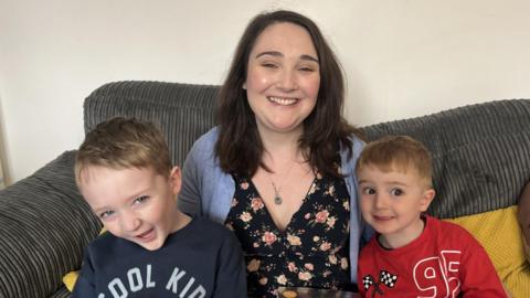 Two young boys and their mum sitting on a grey cord sofa looking at the camera.
On the left is a five year old with brown hair laughing at the camera, in the middle is a woman with long brown hair, wearing a flowery dress and cardigan smiling at the camera, to the right is a little boy with blonde hair wearing a red jumper and is smiling. 