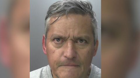Lance Percival, who has short grey hair and a stern expression, wearing a grey t-shirt in a police custody picture.