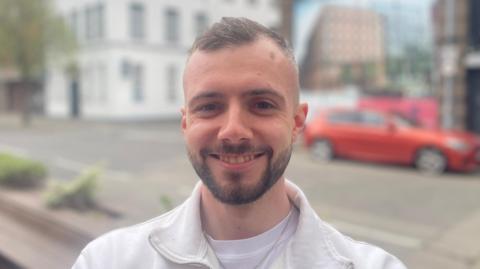 A man with short brown hair and a short brown beard and moustache smiles at the camera. He wears a white quarter zip jumper with a white t-short underneath. 