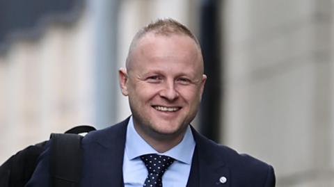 Jamie Bryson is wearing a light blue shirt and a navy and white spotted tie. He is also wearing a navy blazer. He has a black rucksack over one shoulder. The background is blurry. 