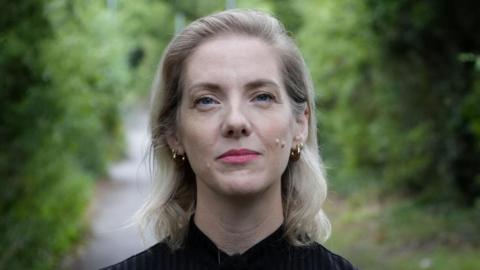 Karen Whybro has shoulder-length blonde hair and is wearing a black top while smiling. She is standing on a path flanked by bushes and trees.