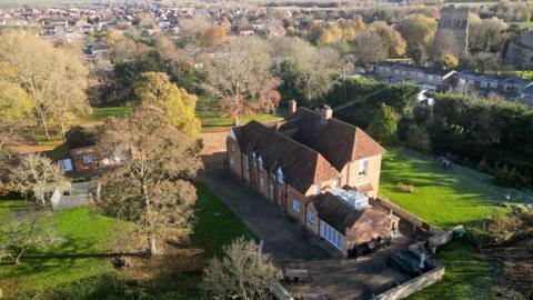 A large detached house surrounded by gardens in Marston Moretaine. There is a church tower and houses in the background. It is an aerial view picture.