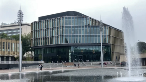 A shiny glass multi storey office building, in a city square with a large fountain in front of it.