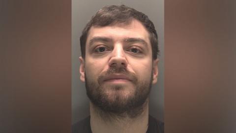 A police mugshot of Lee Harrison, 27, who has brown hair and a brown beard and smirks at the camera.