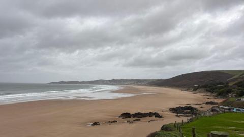 wide expanse of sandy beach and sea with rock clusters, green hills, under a grey, cloudy sky