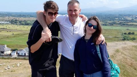 A man puts his arms around a teen boy and girl, while standing on a hill in the countryside.