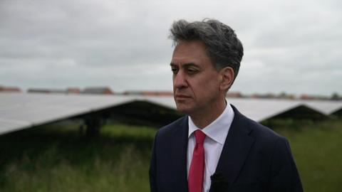 Energy Secretary Ed Miliband standing in front of the Castle Hill Hospital solar farm. He has dark hair and is wearing a white shirt and red tie and a dark suit.