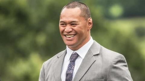 A man looks to the right and smiles. He is wearing a grey jacket, purple tie and white shirt.