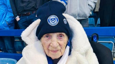 Joan Geary is well wrapped up in a white furry coat, Portsmouth FC hat and club scarf as she sits in the stands at the ground.