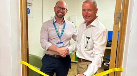 A man wearing a shirt and a blue lanyard shakes the hand of a man wearing a white shirt. The pair are standing in the new suite, with a yellow ribbon seen in front of them.