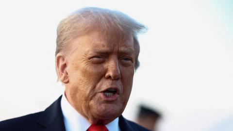 Donald Trump, wearing a red tie and dark suit, speaks toward the camera.