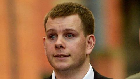 Shaun McKenna has short, brown hair. He is wearing a white shirt and a dark suit jacket. He is staring straight ahead