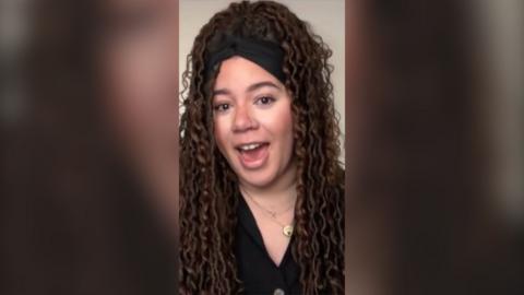 A woman with long brown curly hair in braids talks to the camera. She is wearing a black top and a black hairband.