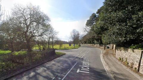 A Google streetview screenshot of Tickenham Road, at the junction of Court Lane. It is a rural road with fields and trees on one side and an old brick wall with large overhanging trees on the right. 