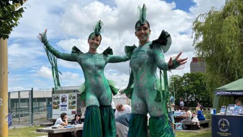 Two women on stilts dressed as green sea creatures. They are holding their arms up and smiling. People are sat on benches behind them. They sky is blue with white clouds. 