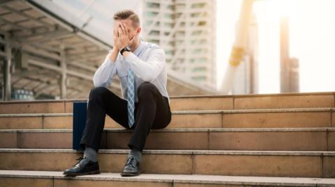A man in office clothes sits on some steps. He looks bereft of hope. 