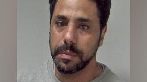 Police mugshot of a man with short dark hair and beard looking towards the camera