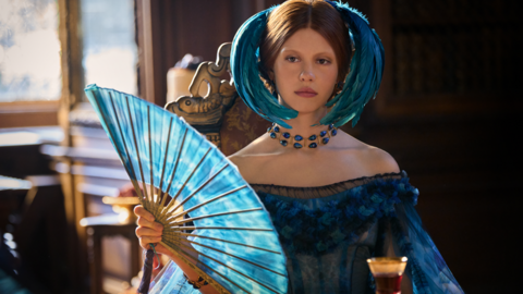 Actress is Mia Goth, who plays Lady Elizabeth Harlander in the film is sitting at a table wearing an elaborate blue headdress and holding a blue fan. She is wearing a blue dress and there is a glass on a table in front of her
