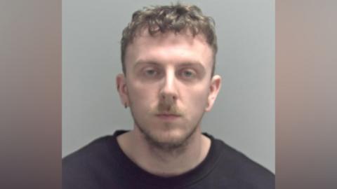 A police mugshot of William Caton, who has short light brown hair, slightly curly at the top of his head. He has a stubbly beard and is wearing a black sweatshirt.
