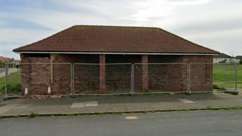 A one-storey brick-built building with a brown tiled roof, surrounded by metal fencing, a pathway and a grassy area