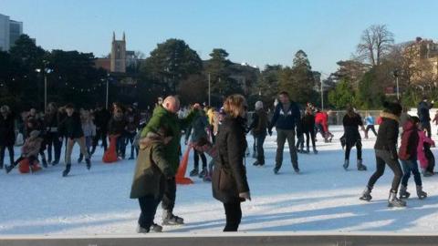 Scores of people wrapped up in coats and hats, skating on an outdoor ice rink on a sunny day. The scene is set against a backdrop of trees with a church tower poking over the tree line in the distance.