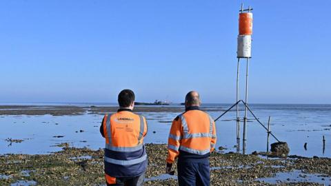 Two men wearing hi-vis orange jackets have their backs to the camera. They look out from a beach to the sea - where there are sand banks and a ship to be used for target practice in the background.