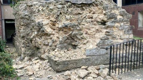 Light coloured bricks are seen damaged and in a pile of rubble, with a black railing and green bushes next to the wall