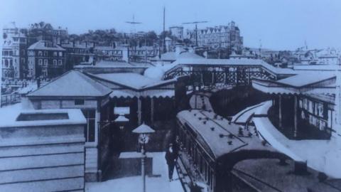  A black and white postcard image of an old train station, with a train stopped at one of the platforms. 