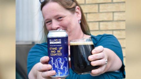 A blonde woman in a teal top smiling at the camera and holding a pint of beer in one hand and a can in the other, with the branding for "Elusive Brewing" and the words "stand up and be counted" written on it. 