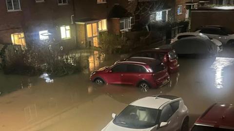 A high shot of a street being flooded by muddy water. Several cars have the water up to the middle of their wheels and the water can be seen up against the bottom of several front doors