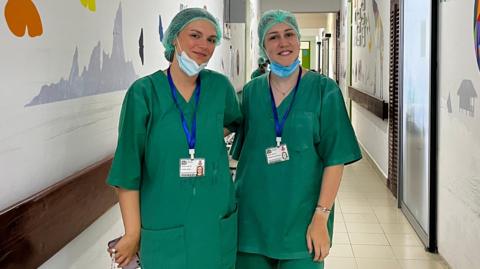 Two women wearing green scrubs with face masks under their chins pictured in a hospital corridor