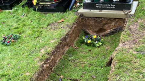 A photograph of the grave with a black headstone and turf which is sunk about 10 centimetres below the ground. Flowers can be seen on the turf.