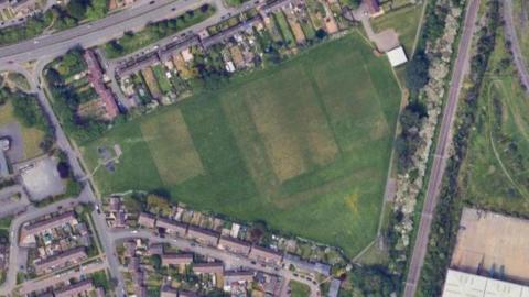 Aerial shot of housing estate with a large triangular playing field. Various sports pitches are marked out on the grass. A small clubhouse is visible in the top right